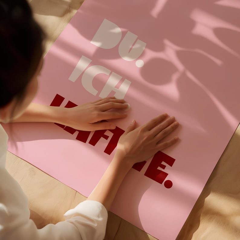 Framed poster with bold German text “Du. Ich. Kaffee.” in playful retro fonts on a solid pink background, leaning against a white wall.
