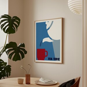 Op de afbeelding: Een ingelijste wandkunstprint met een grafische illustratie van een witte theepot die vloeistof in een effen rode mok schenkt tegen een blauwe achtergrond. De tekst TEA TIME staat in donkerblauwe hoofdletters rechtsonder op het kunstwerk.