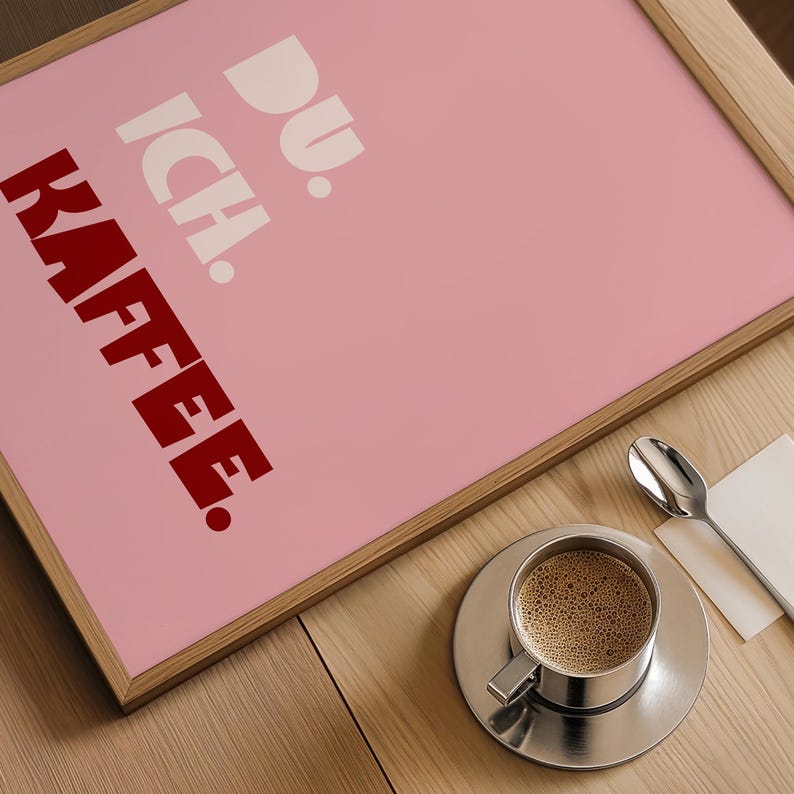 Framed poster with bold German text “Du. Ich. Kaffee.” in playful retro fonts on a solid pink background, leaning against a white wall.