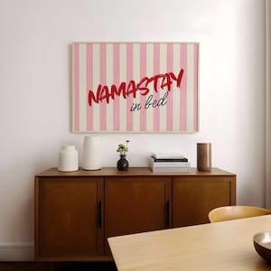 Framed poster with pink and cream vertical stripes, featuring the phrase “Namastay in bed” in bold red and navy handwritten fonts, styled as cozy bedroom wall decor.