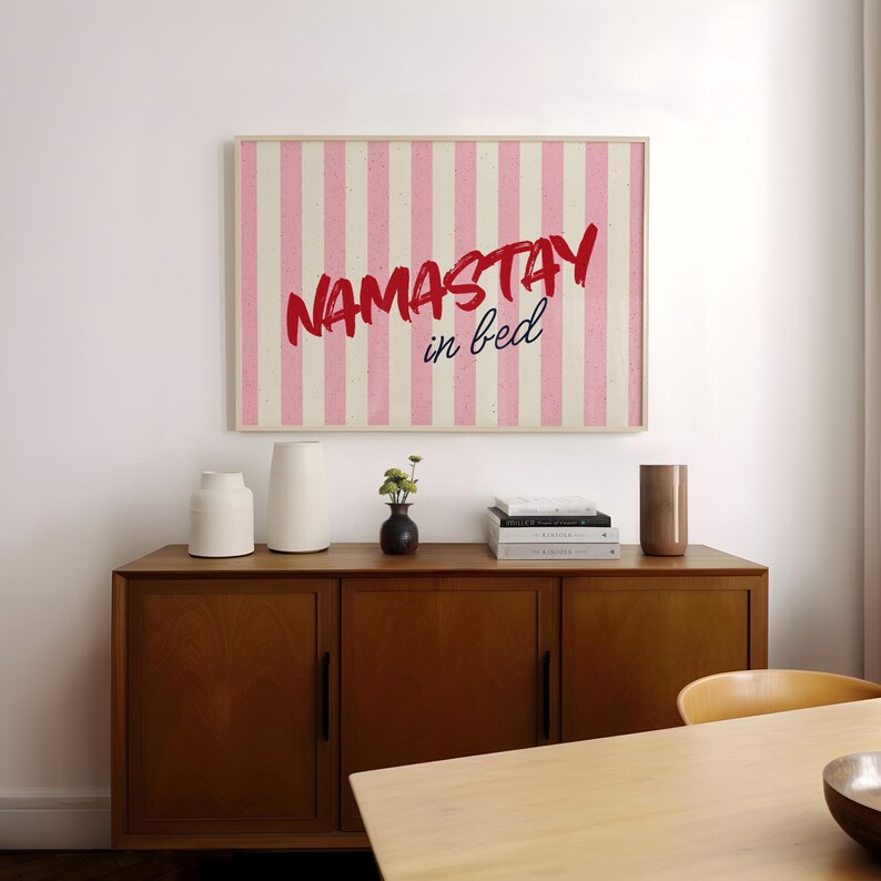 Framed poster with pink and cream vertical stripes, featuring the phrase “Namastay in bed” in bold red and navy handwritten fonts, styled as cozy bedroom wall decor.