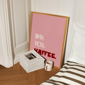 Framed poster with bold German text “Du. Ich. Kaffee.” in playful retro fonts on a solid pink background, leaning against a white wall.