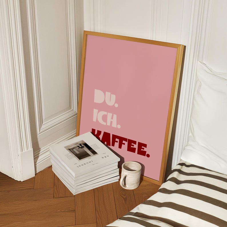 Framed poster with bold German text “Du. Ich. Kaffee.” in playful retro fonts on a solid pink background, leaning against a white wall.