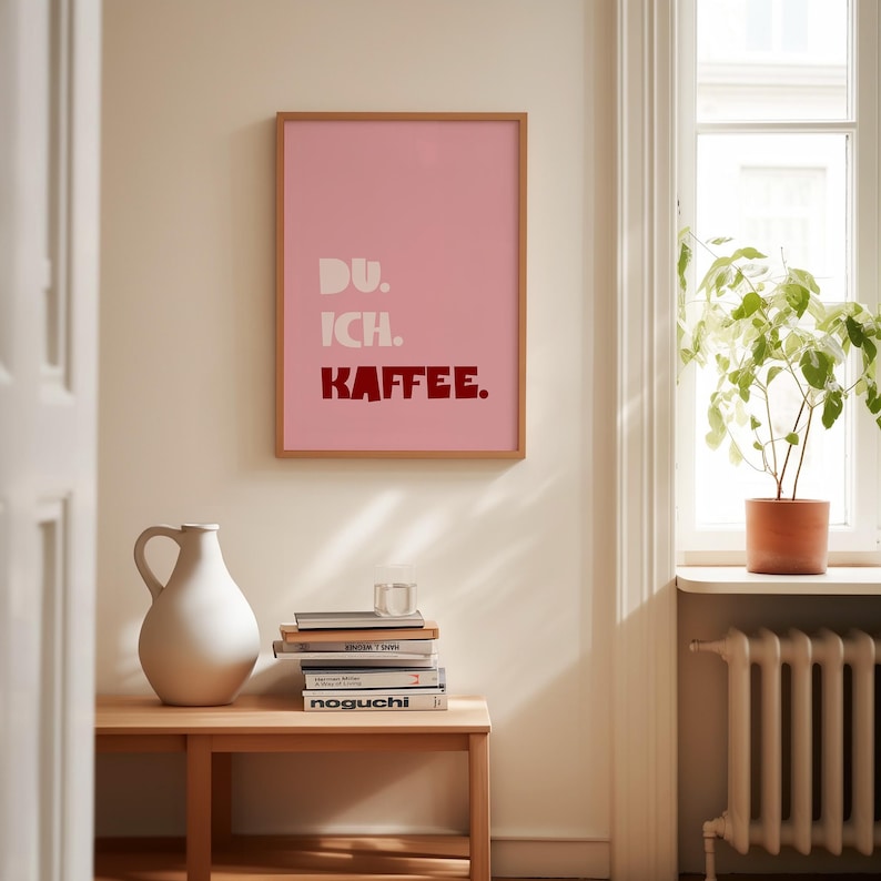 Framed poster with bold German text “Du. Ich. Kaffee.” in playful retro fonts on a solid pink background, leaning against a white wall.