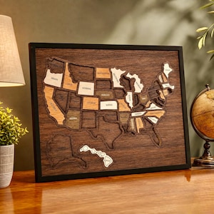 May include: A framed wall art piece featuring a layered wooden map of the United States. The map displays individual states in varying shades of light and dark wood tones, set against a dark brown wood grain background within a black frame.