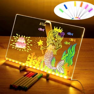 May include: A light-up drawing board with a mermaid drawing on it. The board has a yellow light around the edges and comes with multi-colored markers. The drawing says "Elsa" at the top.