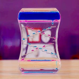 Könnte beinhalten: Ein klarer Flüssigkeits-Bewegungstimer in Sanduhrform mit blauen und rosa Flüssigkeitstropfen. Der obere Bereich ist blau und der untere Bereich ist rosa. Der Timer hat eine klare Flüssigkeit in der Mitte mit kleinen, farbigen Tropfen, die sich langsam bewegen.