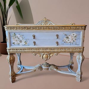 Puede incluir: Una mesa consola antigua pintada en azul claro y dorado con dos cajones y tallas ornamentadas. La mesa presenta diseños florales y una parte superior decorativa.