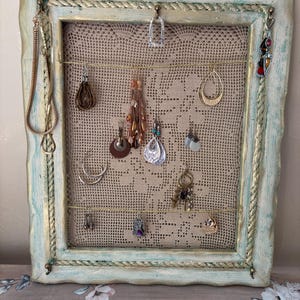 May include: A framed earring holder with a distressed blue and gold finish. The frame holds a beige lace fabric, with various earrings and a necklace hanging from it. The earrings are in different styles and colors.
