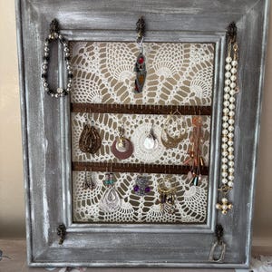 May include: A gray, distressed wooden framed jewelry organizer with a white lace backing. Various earrings, bracelets, and necklaces hang from the frame. The frame has decorative metal corner accents.