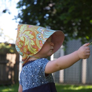 May include: A young child wearing a floral patterned sun hat with a pink brim, pointing with their right hand. The child is wearing a blue and white patterned dress.