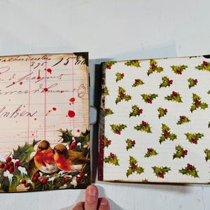 May include: Two open Christmas-themed journals. The left journal features a vintage ledger design with text and red splatters, and a robin and holly illustration. The right journal has a white background with a holly pattern.