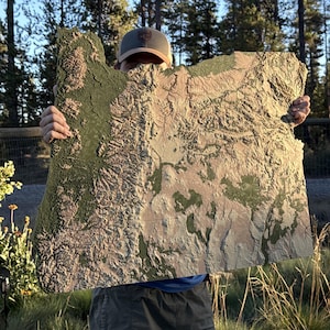 Puede incluir: Un gran mapa en relieve tridimensional de Oregón, sostenido frente a una persona. El mapa presenta características topográficas detalladas en tonos verdes, marrones y beige, con una superficie texturizada. El mapa mide aproximadamente 76 cm de ancho.
