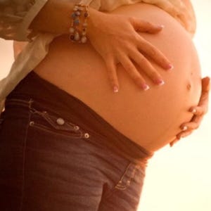 Puede incluir: Primer plano de un vientre embarazado, suavemente sostenido por las manos. Se ve una pulsera en la muñeca. La persona lleva vaqueros azul oscuro. La imagen está bañada en una cálida luz dorada.