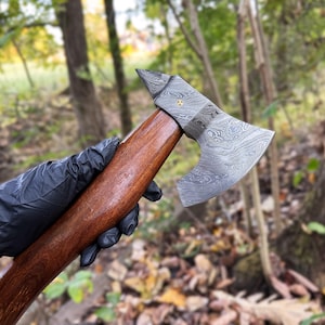 May include: A hand wearing a black glove holds a handmade axe with a wooden handle and a Damascus steel head. The axe has a unique pattern and a sharp blade.