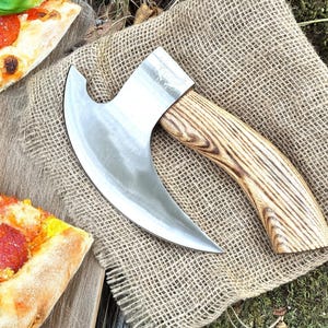 May include: A silver pizza cutter with a wooden handle. The cutter is shaped like an axe and has a curved blade. The handle is made of wood with a dark brown stain.