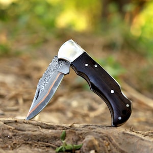May include: A folding pocket knife with a dark wood handle and a silver bolster. The blade has a patterned design with a copper-colored accent. The knife is open, displaying the blade.