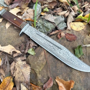 May include: A large, silver,  damascus steel hunting knife with a brown wooden handle. The knife is lying on a bed of brown leaves.