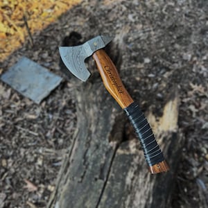 May include: A hand axe with a patterned Damascus steel blade and a wooden handle featuring the engraved name Christopher. The handle includes a black wrapped grip section near the base and a small metal pin detail at the bottom end of the wood.