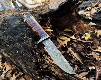 May include: A hunting knife with a patterned steel blade and a brown and black wood handle.