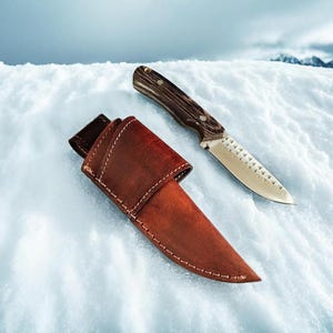 May include: A hunting knife with a wooden handle and a silver blade rests on a snowy surface next to its brown leather sheath. The knife has a serrated edge and a dark-colored handle. The sheath is open.