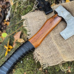 Könnte beinhalten: Eine handgeschmiedete Axt mit einem Holzstiel, der in schwarzes Leder gewickelt ist. Die Axtkopf hat ein Damastmuster und liegt auf einer Jutestoffoberfläche.