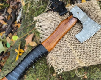 May include: A hand-forged axe with a wooden handle wrapped in black leather. The axe head has a Damascus steel pattern and is resting on a burlap surface.