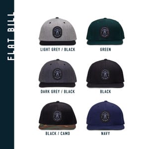 May include: Five different color variations of a flat bill baseball cap with a black and white dog graphic and the text "Lived With A Canine" in a circle. The caps are available in light grey with a black bill, dark grey with a black bill, black with a black bill, black with a camo bill, and navy blue with a navy blue bill.