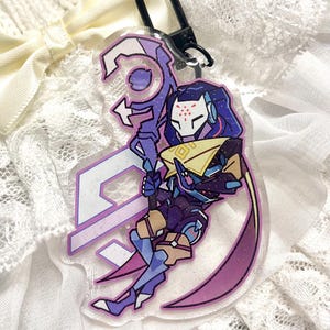 Op de afbeelding: Een heldere acryl sleutelhanger met een paars en blauw karakterontwerp met een wit masker en een staf. De sleutelhanger heeft een zwarte sluiting en een glitterafwerking. Het personage houdt een staf vast.