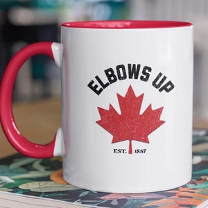 May include: White ceramic mug with a red rim and handle. The mug has a black graphic of a Canadian maple leaf with the text "Elbows Up" above it and "Est. 1867" below it.
