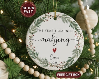 May include: A white ceramic Christmas ornament with the text "THE YEAR I LEARNED mahjong" in black script, a red heart, and the name "Cara" with the year "2025". The ornament is decorated with green leaves and red berries.