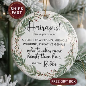 May include: A round ceramic ornament with the word "Hairapist" in black script, surrounded by a green and red floral design. The text defines "Hairapist" and includes the phrase "who touches more hearts than hair."