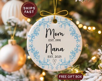 May include: A white ceramic Christmas ornament with blue floral accents and gold string. The ornament reads "Mom EST. 1991" and "Nana EST. 2025" with a heart symbol. The background includes a Christmas tree and ornaments.