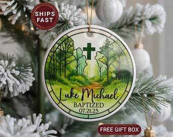 May include: Round ceramic ornament with a forest scene, a green cross, and the names "Luke Michael" with the date "07.21.25" in black script. The ornament hangs from a gold string and is displayed on a Christmas tree.