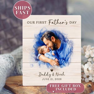 May include: Wooden plaque with a watercolor image of a father and baby. The text reads "OUR FIRST Father's DAY", "Daddy & Noah", and "JUNE 21, 2026". The plaque has a light wood grain background and is surrounded by a blue watercolor effect.