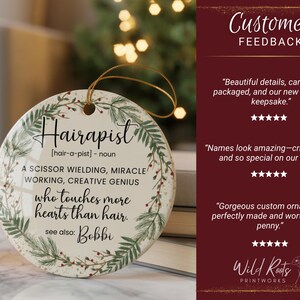 May include: A round, white ceramic ornament with the word "Hairapist" in elegant script, surrounded by a green and red floral wreath. The ornament includes text defining "Hairapist" and a gold string for hanging.