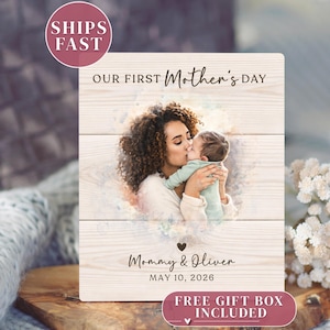 May include: A wooden plaque with a photo of a mother kissing a baby. The text reads "OUR FIRST Mother's DAY", "Mommy & Oliver", and "MAY 10, 2026". A heart graphic is below the names. The plaque is light-colored with a wood grain pattern.