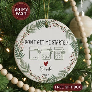 May include: A round, white ceramic Christmas ornament with the text "DON'T GET ME STARTED" and three jar illustrations. The ornament includes the name "Sarah" and the year "2025". It is decorated with a green and red floral border and a gold hanging string.