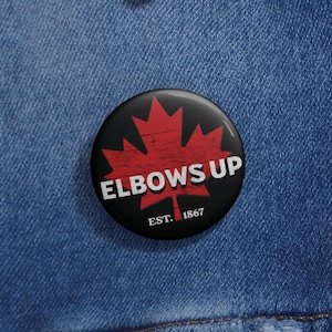 May include: A black and red button featuring a red maple leaf and the text "Elbows Up" in white. Below the text is "EST. 1867" in white.