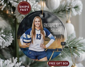 May include: A round ceramic ornament featuring a volleyball player in a blue and white uniform holding a volleyball. The ornament has the name "Sophia" and the year "2025" printed on it. The ornament is hanging on a Christmas tree.