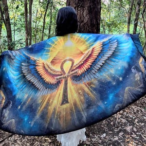 Puede incluir: Un vibrante chal de temática celestial con un símbolo ankh central con alas extendidas, sobre un fondo de pirámide y cielo estrellado. La paleta de colores incluye azules, dorados y naranjas, con ilustraciones detalladas de figuras angelicales.