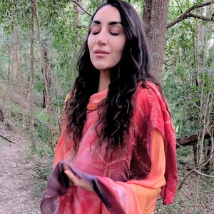 May include: A woman wearing a red and orange scarf with script text. The scarf drapes over the shoulders. The background is a forest with trees and foliage.