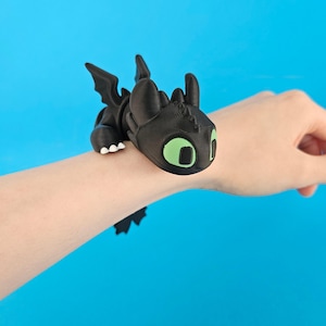Op de afbeelding: Een zwart drakenarmband met groene ogen en witte tanden. De draak heeft vleugels en rust op de arm van een persoon. De achtergrond is effen blauw.