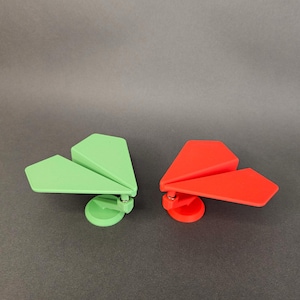 Pode incluir: Dois objetos decorativos em forma de avião de papel. Um é verde claro, o outro é vermelho brilhante. Cada um tem uma base circular e uma conexão articulada. As asas estão anguladas para cima. O fundo é cinza liso.