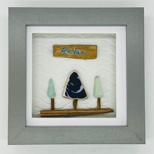 May include: A framed artwork featuring a serene scene. The artwork includes three stylized trees, a central tree with a dark blue and white design, and two light green trees. The word "Relax" is written in blue on a wooden sign.