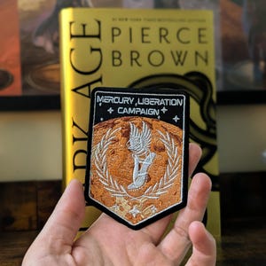 May include: A black and orange embroidered patch with the text "Mercury Liberation Campaign" and a winged figure in the center.