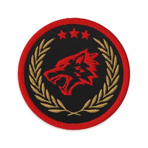Op de afbeelding: Geborduurde patch met een rode wolfskop op een zwarte achtergrond, omringd door een gouden lauwerkrans en een rode rand. Het ontwerp bevat drie sterren boven de wolfskop.