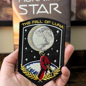 May include: Black embroidered patch with the text "THE FALL OF LUNA" and a detailed moon design. The patch features a red and black figure, stars, and a gold laurel wreath. The background is a book titled "MORNING STAR".