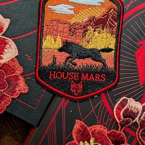 May include: Embroidered patch with a landscape scene featuring a black wolf leaping across a field, with a sunset sky and mountains. The patch has the text "HOUSE MARS" in red. The patch is on a black surface with red floral embroidery.