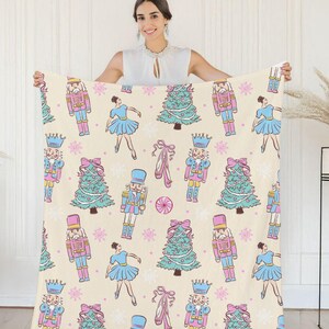 May include: A cream-colored blanket featuring a repeating pattern of Christmas elements. The design includes nutcrackers, ballerinas, Christmas trees, ballet slippers, and snowflakes in shades of pink, blue, and green.